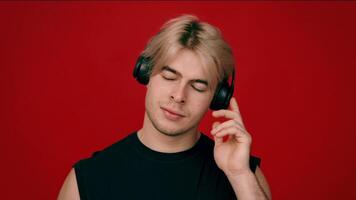 Studio portrait of young blonde guy listening to music in headphones and dancing, red background, slow motion video
