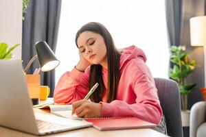 Tired teenage girl doing homework and writing in notebook at home photo