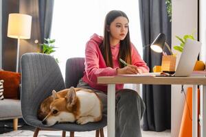 Focused female student taking notes while studying online with dog sleeping nearby photo