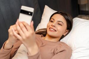 Happy young girl, teenager lying in bed using smartphone at night, text message photo
