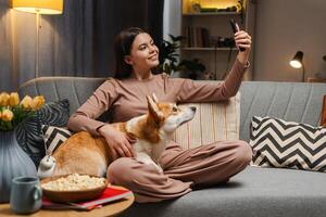 Smiling girl relaxing on sofa with corgi and taking selfie with smartphone photo