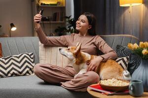 Smiling girl relaxing on sofa with corgi and taking selfie with smartphone photo