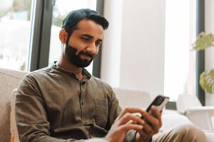 Smiling bearded Indian man holding mobile phone using mobile app, sitting on comfortable sofa photo