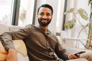 Happy, smiling Indian man relaxing on comfortable sofa using smartphone looing at camera photo