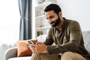 Happy, smiling Indian man using smartphone browsing social media at home on sofa photo