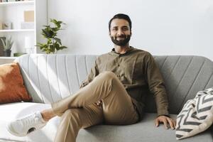 Confident entrepreneur, smiling Indian man sitting on sofa at home, looking at camera photo
