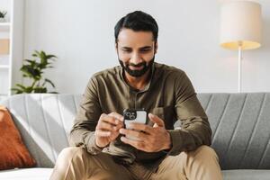 Smiling Indian man sitting on sofa holding mobile phone, using mobile app at home, text message photo