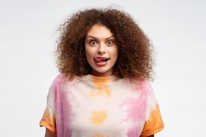 contento joven mujer con Rizado pelo vistiendo casual verano t camisa demostración lengua, mirando a cámara foto