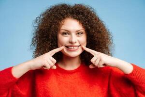 Attractive smiling plus size model with curly hair pointing fingers at teeth, looking at camera photo