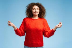 Smiling attractive woman with curly hair meditating with closed eyes isolated on blue background photo