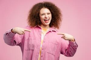 alegre sonriente mujer con Rizado pelo señalando dedos a sí misma mientras mirando a cámara foto