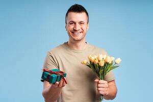 Handsome smiling young man looking at camera, holding bouquet of flowers and gift in hand photo