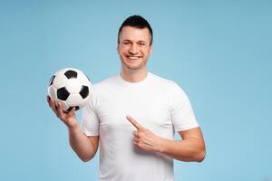 Cheerful football fan holding ball and pointing at it on blue background photo