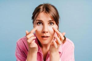 Beautiful woman with patches under eyes looking in mirror, posing isolated on blue background photo