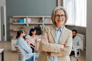 Senior businesswoman smiling with crossed arms in busy office looking at camera photo