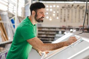 Happy young bearded man choosing new window frame in in the construction department photo