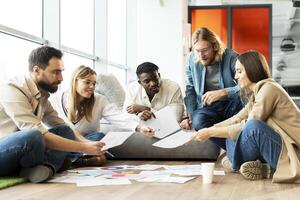 Group multiracial creative designers discussing documents while sitting on floor in modern office photo