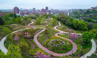 A close-up of a city park seen from above, with winding paths, blooming flowers, and the cityscape in the background photo