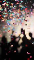 A close-up of falling confetti in focus, with blurred silhouettes of people celebrating in the background photo