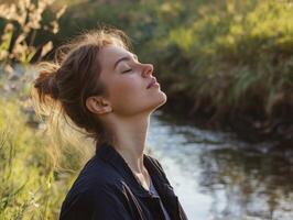 Woman outdoors, standing by a stream with her eyes closed, head tilted back, breathing deeply, with space for copy in the peaceful landscape photo