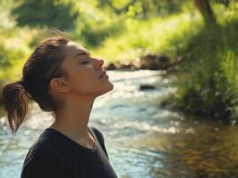 Woman outdoors, standing by a stream with her eyes closed, head tilted back, breathing deeply, with space for copy in the peaceful landscape photo