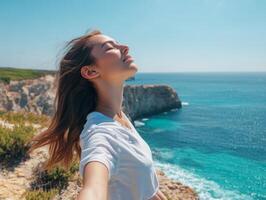 A woman enjoying the fresh breeze on a cliffside overlooking the ocean, eyes closed, arms wide open, with clear sky and ocean view for copy space photo