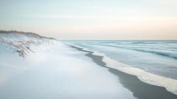 A calm winter beach with snow on the dunes and icy waves lapping at the shore, under a pale blue sky photo