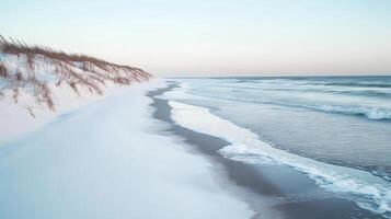 A calm winter beach with snow on the dunes and icy waves lapping at the shore, under a pale blue sky photo