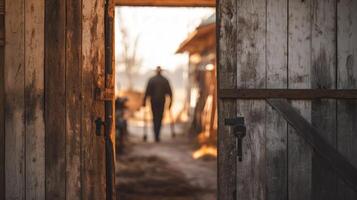 A rustic barn door slightly open in the foreground, with a blurred farmer walking inside with farming tools in the background photo