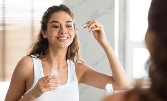 facial protección de la piel. atractivo hembra aplicando suero en cara hidratante y cuidando para piel en pie cerca espejo en moderno baño adentro. belleza rutina. piel cuidado concepto. selectivo atención foto