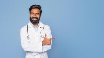A cheerful Indian doctor with a dark beard and a stethoscope around his neck is pointing to his left while smiling. He is wearing a white coat and stands against a solid blue background, copy space photo