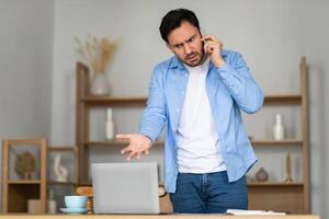 A man stands in front of his laptop in a home office, expressing frustration and confusion during a phone call. His body language, with one hand on his hip and a furrowed brow photo