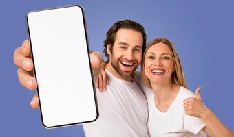 A happy couple stands in front of a blue background, holding a smartphone with a blank screen in front of them. The man is smiling and pointing at the phone, while the woman is giving a thumbs up photo