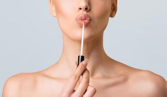 A close-up image of a woman applying lip gloss to her lips with a wand applicator. She is looking directly at the camera and the image is cropped to focus on her face and upper chest. photo