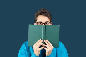 A young man sits against a solid dark blue background, holding an open book in front of his face. He wears glasses and a bright blue sweater, peering over the top of the book photo