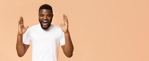 A young black man wearing a white t-shirt smiles and looks shocked while raising his hands in the air. He has a light brown complexion and a full beard. He is standing in front of a peach background. photo