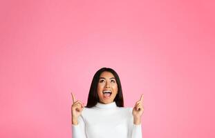 A young Asian woman with long dark hair is pointing upwards at copy space with both hands, looking up with a wide smile and an excited expression. She is against a solid pink background. photo