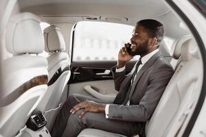African American businessman in a suit is sitting in the backseat of a car, talking on his phone with a smile on his face. He is dressed in a gray suit, white shirt, and a dark tie photo