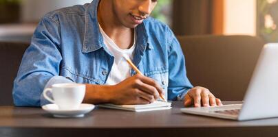 Cropped of guy is seated at a table with a laptop open in front of him. He is holding a cup of coffee, appearing focused on his work or task at hand in a casual setting. photo