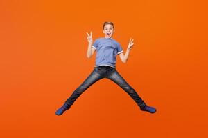 Emotional carefree boy jumping and showing peace gesture on orange studio background, empty space photo