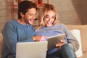 Smiling Man And Woman Using Laptop And Digital Tablet Websurfing And Chatting In Social Media Sitting On Sofa Indoor photo