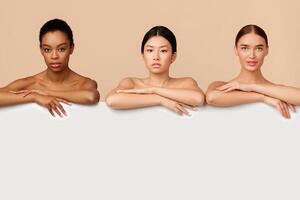 Three Multiethnic Ladies Standing Naked Posing Behind White Board For Text On Gray Background In Studio. Empty Space photo