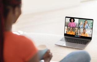 A woman sits on a couch with a laptop in front of her, attending a virtual meeting with four other people on the screen. The laptop is open and displaying a call with four participants photo