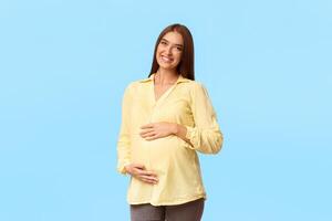 Mother-To-Be. Happy Pregnant Lady Smiling To Camera Posing Touching Belly Standing Over White Background In Studio photo