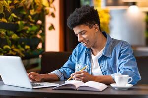 A smart African American young guy is seated at a table, working on a laptop while sipping from a cup of coffee. He appears focused and engaged in his work. photo