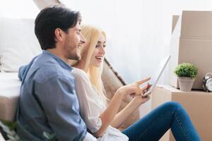 Cheerful Couple Using Tablet Computer Sitting Among Moving Boxes After Relocation To New Place. Empty Space photo