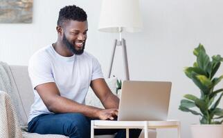 A smiling Black man with a beard sits on a couch, working on a laptop. He is wearing a white t-shirt and blue jeans. There is a floor lamp behind him and a plant in a white pot to the right photo