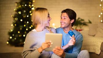 Couple buying Christmas gifts online, using tablet and credit card, sitting in front of Christmas tree photo