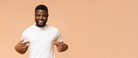 A happy black man with short dark hair, a beard, and a white T-shirt is smiling and pointing at himself with both hands. He is standing in front of a peach background, copy space photo