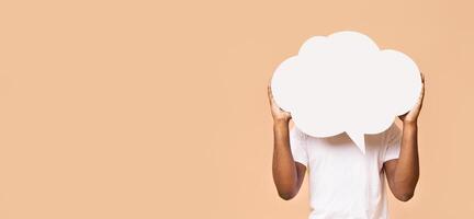 A person in a white t-shirt holds a blank, cloud-shaped speech bubble in front of their face. The background is a light peach color, panorama with copy space photo